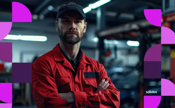 Homem com barba e boné preto, vestindo um macacão vermelho de trabalho, posa com os braços cruzados em uma oficina ou ambiente industrial ao fundo desfocado. Nas laterais da imagem, há grafismos geométricos em tons de roxo e rosa com o logotipo da Sólides no canto inferior direito.